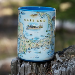 May include: A blue ceramic mug with a map of Cape Cod printed on it. The map shows the coastline, towns, and landmarks. The mug has a blue rim and handle.