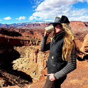 May include: A person wearing a black vest and a black and white striped shirt is standing on a cliff overlooking a canyon. They are holding a white and yellow travel mug with the text "Rooster Coffee" on it.