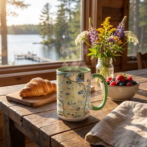 May include: A ceramic mug featuring a detailed map illustration of Wisconsin with a green interior and handle, placed on a wooden table next to a croissant on a wooden board, a bowl of fresh berries, and a glass vase filled with colorful wildflowers.