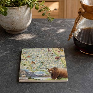 May include: A square coaster featuring a detailed illustrated map of Glacier National Park. The design includes topographical terrain, a large brown bear in the foreground, various wildlife icons, and a red tour bus. The text reads Glacier National Park.