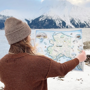 Puede incluir: Una persona sosteniendo un mapa de Alaska, con la palabra "ALASKA" en un lugar destacado. El mapa presenta ilustraciones de vida silvestre y puntos de referencia. La persona lleva un gorro y un suéter de punto marrón, con una cordillera nevada al fondo.