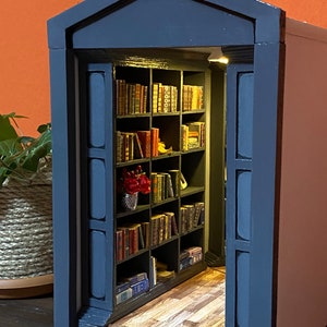 Może przedstawiać: Miniaturowa drewniana biblioteczka z ciemnoniebieskim wykończeniem i drewniana podłogą. Biblioteczka jest wypełniona miniaturowymi książkami i małym bukietem czerwonych kwiat&oacute;w.