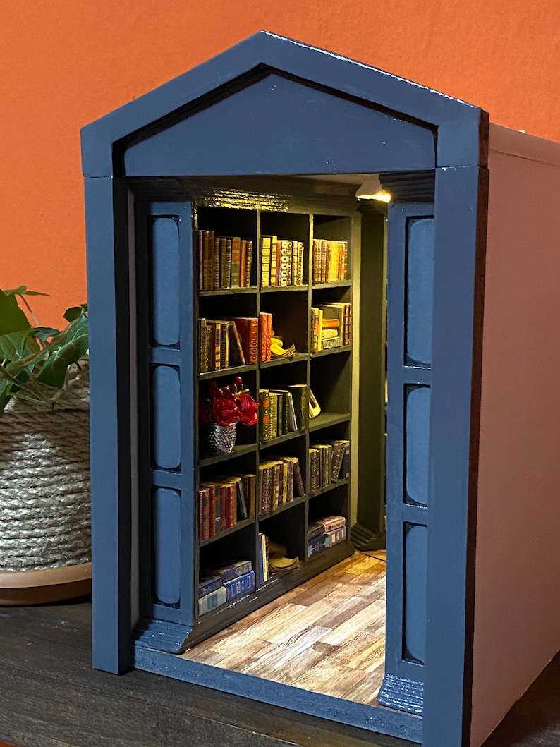 Może przedstawiać: Miniaturowa drewniana biblioteczka z ciemnoniebieskim wykończeniem i drewniana podłogą. Biblioteczka jest wypełniona miniaturowymi książkami i małym bukietem czerwonych kwiat&oacute;w.