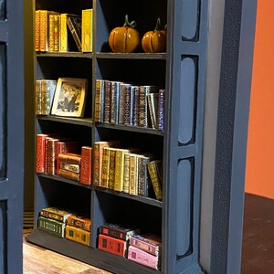 Może przedstawiać: Miniaturowa biblioteczka z pięcioma p&oacute;łkami wypełnionymi miniaturowymi książkami. Biblioteczka jest pomalowana na czarno i ma ciemnoszary obramowanie. Na drugiej p&oacute;łce znajdują się dwie małe pomarańczowe dynie.