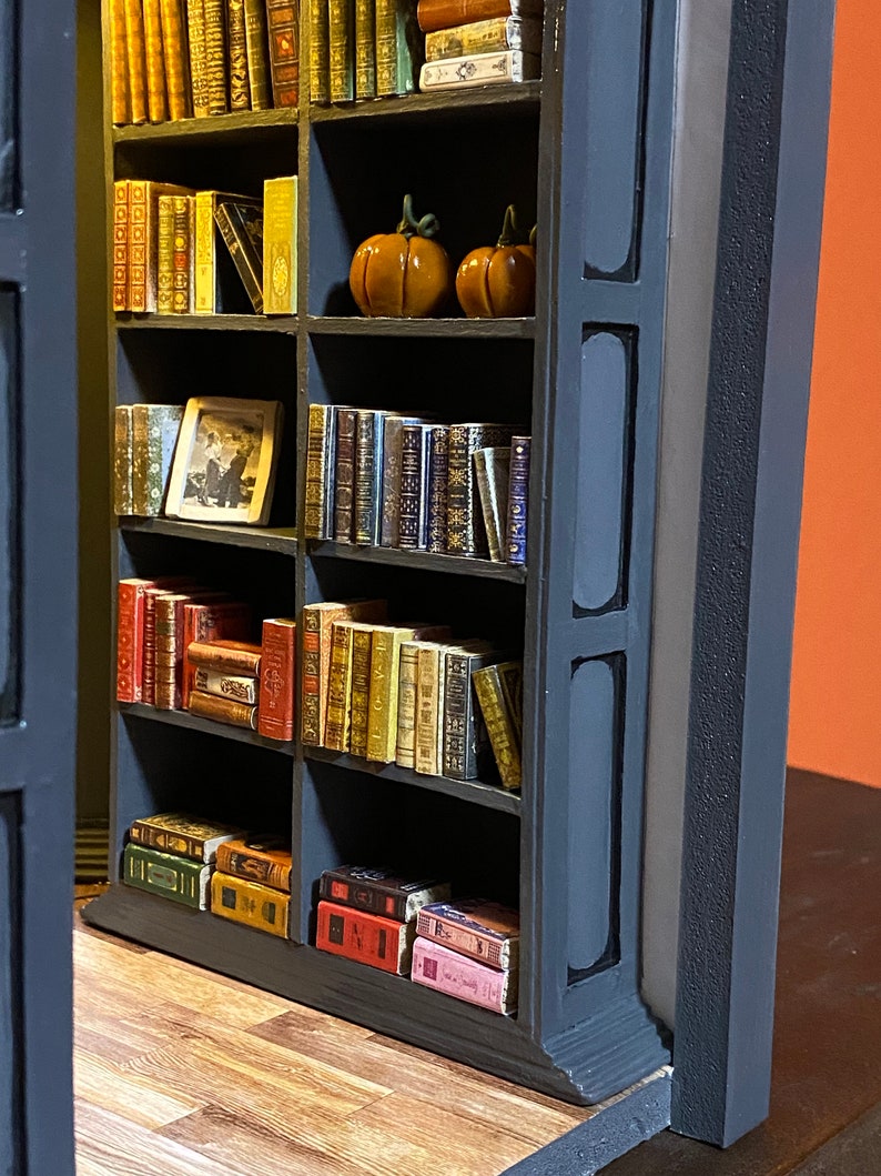 Może przedstawiać: Miniaturowa biblioteczka z pięcioma p&oacute;łkami wypełnionymi miniaturowymi książkami. Biblioteczka jest pomalowana na czarno i ma ciemnoszary obramowanie. Na drugiej p&oacute;łce znajdują się dwie małe pomarańczowe dynie.
