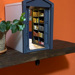 Może przedstawiać: Miniaturowa drewniana biblioteczka z ciemnoszarymi drzwiami i jasnoszarą ścianą z tyłu. Biblioteczka jest wypełniona miniaturowymi książkami i ma kilka małych pomarańczowych dyni na g&oacute;rnej p&oacute;łce.