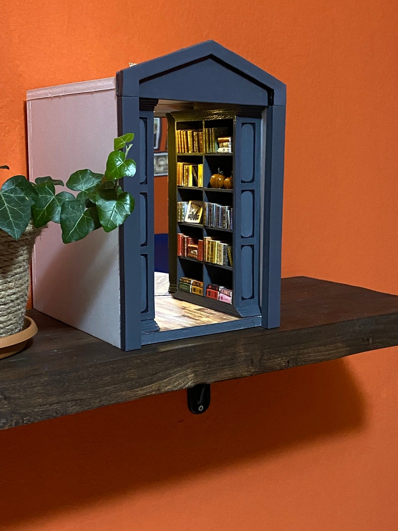 Może przedstawiać: Miniaturowa drewniana biblioteczka z ciemnoszarymi drzwiami i jasnoszarą ścianą z tyłu. Biblioteczka jest wypełniona miniaturowymi książkami i ma kilka małych pomarańczowych dyni na g&oacute;rnej p&oacute;łce.