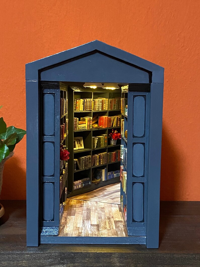Może przedstawiać: Drewniana biblioteczka w ciemnoniebieskim kolorze z sekretnymi drzwiami, kt&oacute;re otwierają się na miniaturową bibliotekę z regałami pełnymi książek. Biblioteczka ma ozdobny, łukowaty szczyt i drewnianą podłogę.