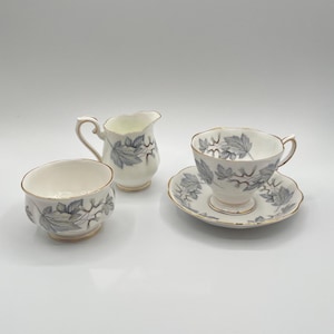 Könnte beinhalten: Ein Vintage-Teeservice mit einer Teetasse, Untertasse, Milchkanne und Zuckerdose. Die weißen Porzellanstücke sind mit einem grauen Blättermuster und goldenen Verzierungen versehen. Die Teetasse steht auf einer passenden Untertasse.