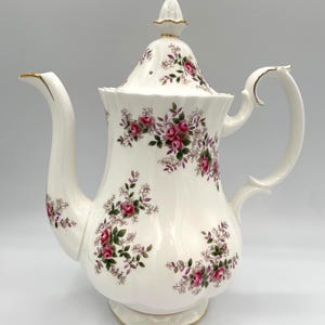Può includere: Caffettiera in porcellana bianca con coperchio, ornata da motivi floreali di rose rosa e finiture dorate. Il manico e il beccuccio sono curvi e la caffettiera poggia su una piccola base smerlata. Un pezzo decorativo.