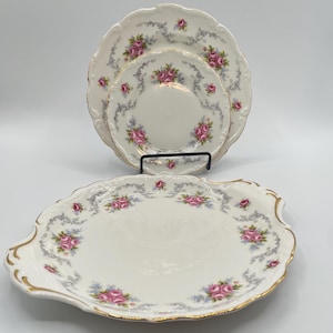 Op de afbeelding: Een set vintage porseleinen servies met een bloemenrozenpatroon. De set bevat drie borden van verschillende formaten en een serveerschaal. De randen zijn geschulpt en afgewerkt met goud. De borden zijn wit met roze rozen en grijze accenten.