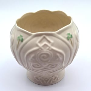 May include: Cream-coloured ceramic bowl with a scalloped rim and a pedestal base. The bowl features embossed Celtic knot designs and green shamrock accents. The bowl is approximately 13 cm in diameter and 10 cm tall.