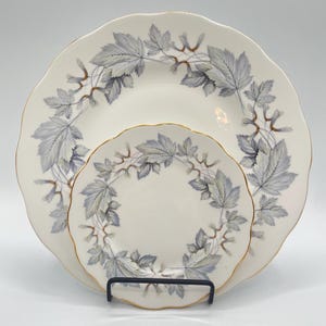 Könnte beinhalten: Ein Set aus drei weißen Porzellantellern mit einem zarten Blatt- und Samenkapsel-Design in Grau- und Brauntönen. Die Teller haben gewellte Ränder und goldfarbene Verzierungen, was eine elegante Ästhetik schafft. Die Teller werden auf einem schwarzen Metallständer präsentiert.
