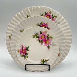 Op de afbeelding: Set van drie witte porseleinen borden met een geschulpte rand en gouden rand. Elk bord heeft een bloemenontwerp met roze bloemen en groene bladeren. De borden worden gepresenteerd op een zwarte metalen standaard.