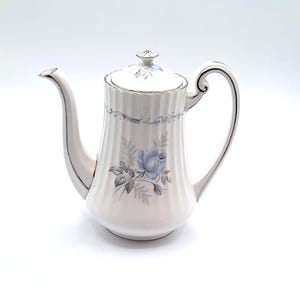 Peut inclure: Cafetière en céramique blanche avec un motif floral de rose bleue. Le pot a un bec verseur incurvé, une anse et un couvercle. Des bordures argentées ornent les bords et la poignée. Une pièce décorative pour servir le café ou le thé.