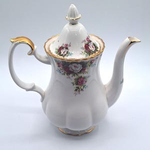 Może przedstawiać: Biały porcelanowy imbryk o falistym korpusie z pasującą pokrywką zakończoną szpiczastym uchwytem. Imbryk ozdobiony jest kwiatowym wzorem w różowe i czerwone róże, podkreślonym złotymi wykończeniami na uchwycie, wylewce, pokrywce i podstawie.