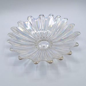 Op de afbeelding: Iriserende glazen kom met een madeliefachtig ontwerp. De kom heeft een centrale, cirkelvormige inkeping en uitstralende, bloembladachtige randen. Het glas heeft een glinsterende, opalescerende kwaliteit en reflecteert licht in verschillende kleuren. Een decoratief stuk.