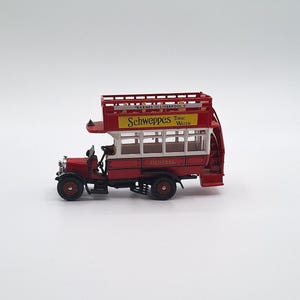 Pode incluir: Um modelo de autocarro de dois andares vermelho com o texto "Schweppes Tonic Water" na lateral. O autocarro de brinquedo tem uma escada na parte de trás e a palavra "General" está impressa na lateral. O autocarro tem rodas pretas.