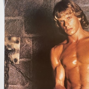 May include: A shirtless man with long blond hair sits in a dark room with a brick wall. He is wearing a leather bracelet on his wrist.