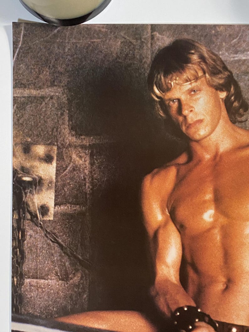 May include: A shirtless man with long blond hair sits in a dark room with a brick wall. He is wearing a leather bracelet on his wrist.