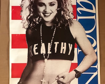 Può includere: Un poster in bianco e nero con una donna con i capelli biondi ricci che indossa una canotta nera con la scritta "HEALTHY" stampata. Indossa una catena d'argento con un ciondolo a forma di croce e un orologio al polso sinistro. Lo sfondo è un motivo a strisce rosso, bianco e blu con la parola "MADONNA" stampata in blu sul lato destro.