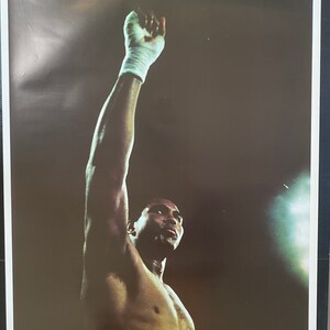 May include: A black and white poster featuring a boxer with his arm raised in victory. The poster has the text "THE GREATEST" in yellow letters at the top.