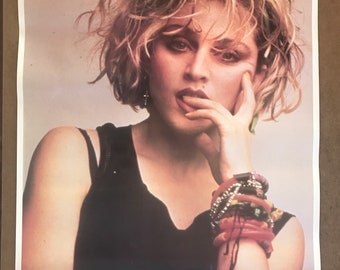 Può includere: Un poster vintage con un ritratto ravvicinato di Madonna. Ha i capelli corti e biondi con foglie verdi al suo interno. Indossa una canottiera nera e diversi braccialetti al polso. Il poster ha la parola "MADONNA" stampata in blu in basso.