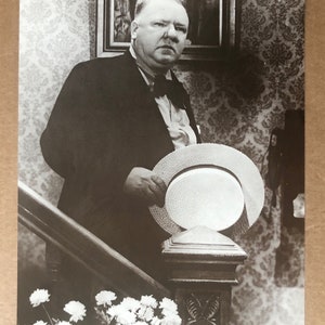 Peut inclure: Photographie en noir et blanc d'un homme en costume et nœud papillon, tenant un chapeau de paille. Il se tient sur un escalier avec un poteau en bois décoratif. Des fleurs blanches sont au bas de l'escalier.