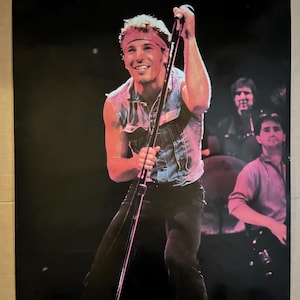 Puede incluir: Un póster de concierto en blanco y negro con un hombre cantando en un micrófono. Lleva un chaleco vaquero, una bandana y pantalones negros. El fondo es oscuro y borroso.