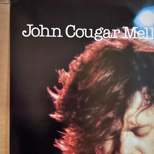 Op de afbeelding: Een zwart-wit concertposter met John Cougar Mellencamp. De poster toont een close-up van Mellencamps gezicht met zijn haar dat een deel van zijn gezicht bedekt. De poster heeft een witte achtergrond met zwarte tekst.