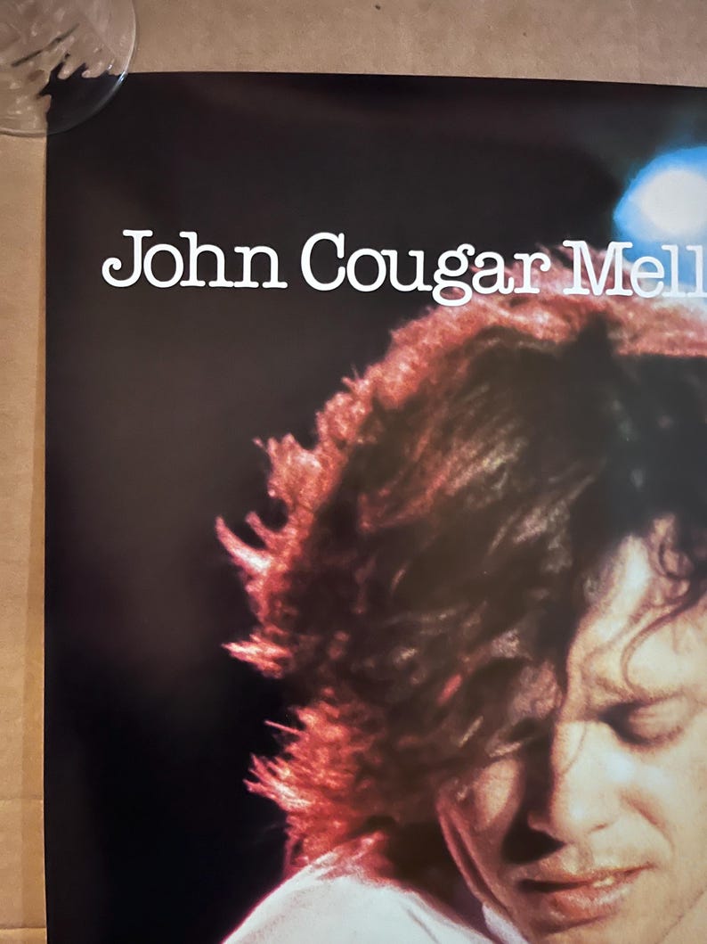 Op de afbeelding: Een zwart-wit concertposter met John Cougar Mellencamp. De poster toont een close-up van Mellencamps gezicht met zijn haar dat een deel van zijn gezicht bedekt. De poster heeft een witte achtergrond met zwarte tekst.