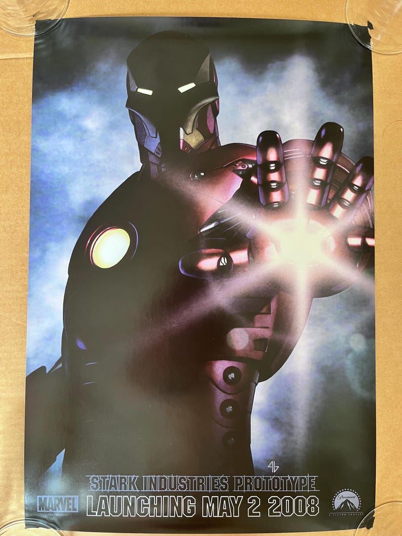 Op de afbeelding: Een rood en zwart Iron Man-pak met een gloeiend licht in de palm van de hand. De tekst "STARK INDUSTRIES PROTOTYPE LAUNCHING MAY 2 2008" staat onderaan de poster.