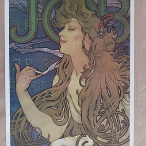 May include: Art Nouveau poster featuring a woman with flowing brown hair, smoking a cigarette. The word "JOB" is prominently displayed in green, stylized lettering. The background is a mix of blue and green with decorative patterns. The poster is framed.