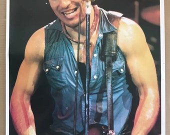 Könnte beinhalten: Ein Poster mit einem Musiker, der live Gitarre spielt. Der Musiker trägt ein rotes Stirnband und ein blaues ärmelloses Hemd. Das Bild wurde während eines Konzerts aufgenommen, mit einem Mikrofon und einem Schlagzeug im Hintergrund.