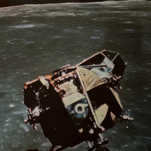 May include: A black and white lunar module with a round window, against a backdrop of the moon's surface and a blue and white Earth in the distance.
