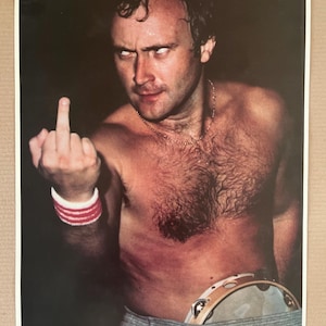 May include: A black and white photograph of Phil Collins giving the middle finger. He is shirtless and wearing a red and white wristband. He is holding a tambourine in his right hand. The photo is captioned "Phil Collins".