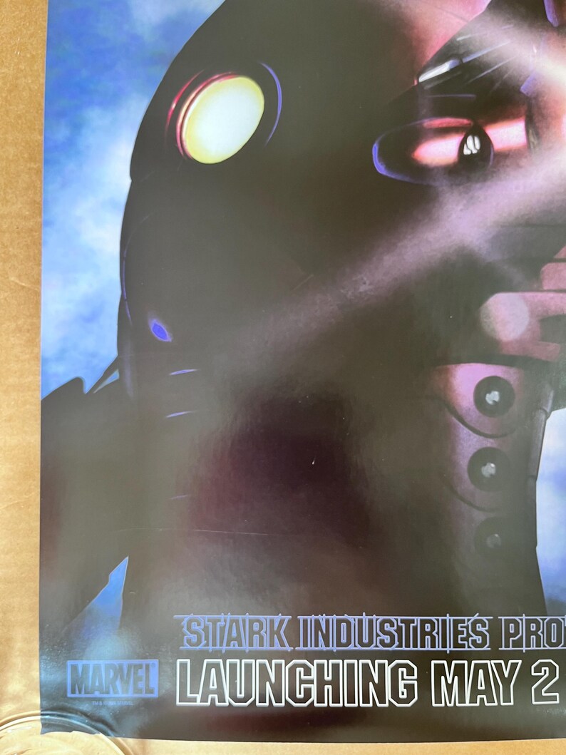 Op de afbeelding: Een zwart en zilveren poster met een futuristisch robotontwerp. De poster heeft de tekst "STARK INDUSTRIES PRO... LAUNCHING MAY 2" onderaan.