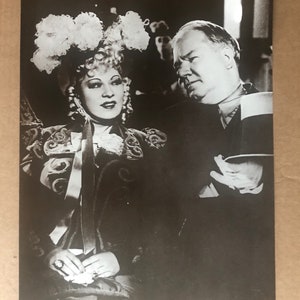 Peut inclure: Une photographie en noir et blanc d'une femme portant un grand chapeau blanc et d'un homme en costume. La femme est assise et l'homme est debout. La femme regarde l'appareil photo et l'homme regarde sur le côté.