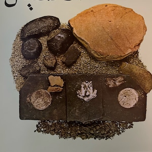 May include: A close-up of a collection of dark brown and black stones, including a large flat stone with a white emblem. The stones are arranged on a light brown surface with a light green background. The Arabic word "الحشيش" is written in black at the top of the image.