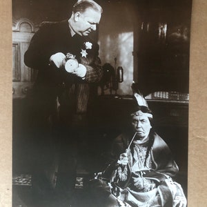 Peut inclure: Une photographie en noir et blanc d'un homme en costume et cravate debout à côté d'un homme amérindien portant une coiffe et des vêtements traditionnels. L'homme en costume tient un petit objet dans sa main.