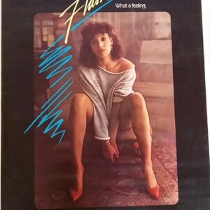 May include: A movie poster for the film Flashdance, featuring Jennifer Beals in a grey sweater and red shoes. The poster has the title "Flashdance" in large, stylized letters and the tagline "What a feeling." The poster also includes the text "Take your passion and make it happen!"