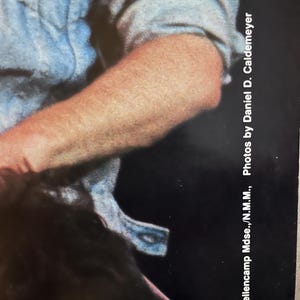 Op de afbeelding: Een close-up van een arm die een lichtblauw overhemd met knopen draagt. De afbeelding is bij de elleboog geknipt en het overhemd is licht gekreukt. De tekst "1984 Mellencamp Muse...N.M.M...Photos by Daniel D. Caldemeyer" is zichtbaar in de rechterbenedenhoek van de afbeelding.