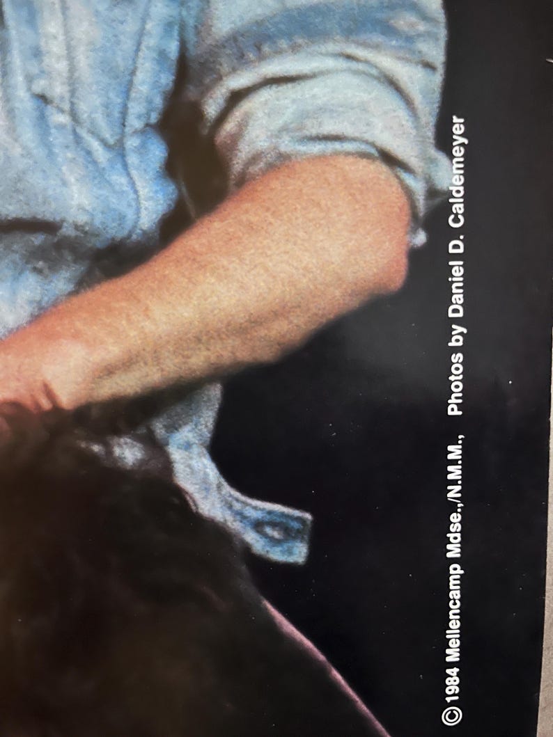 Op de afbeelding: Een close-up van een arm die een lichtblauw overhemd met knopen draagt. De afbeelding is bij de elleboog geknipt en het overhemd is licht gekreukt. De tekst "1984 Mellencamp Muse...N.M.M...Photos by Daniel D. Caldemeyer" is zichtbaar in de rechterbenedenhoek van de afbeelding.