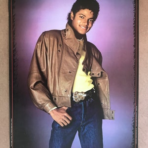 Op de afbeelding: Een vintage poster met een portret van Michael Jackson die een bruin leren jack, een geel shirt, een blauwe jeans en een riem met een grote gesp draagt. De achtergrond is een paarse gradiënt.