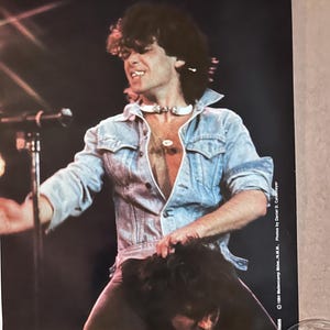 Op de afbeelding: Een zwart-witfoto van een man in een spijkerjack met een witte choker, die op de schouders van een andere man staat. De foto is van een concert en heeft de tekst "1984  Monsters of Rock - N.J. - N.Y.  Photos by Daniel D. Coddington" in de rechterbenedenhoek.