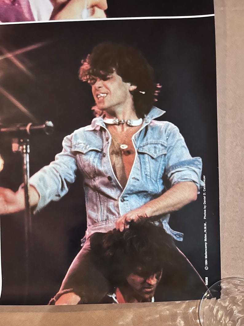 Op de afbeelding: Een zwart-witfoto van een man in een spijkerjack met een witte choker, die op de schouders van een andere man staat. De foto is van een concert en heeft de tekst "1984  Monsters of Rock - N.J. - N.Y.  Photos by Daniel D. Coddington" in de rechterbenedenhoek.