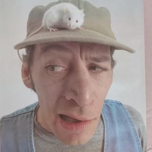 Puede incluir: Un póster que muestra a un hombre con una gorra verde con un ratón blanco encima. El hombre tiene una expresión de sorpresa. El texto en la parte superior dice: "Vern! Just when you think you've won the rat race, along comes a faster rat. KnowWhatImean?"