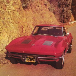 May include: A red 1960s Chevrolet Corvette sports car with a black interior. The car has a California license plate that reads "ZEW 720". The car is parked on a tarmac road with a rocky hillside in the background.