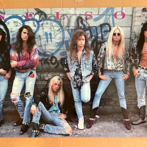 May include: A poster featuring the band Nelson, with six members posing in front of a graffiti-covered brick wall. The band members are wearing denim jeans, jackets, and shirts. The band's name is written in large letters at the top of the poster.
