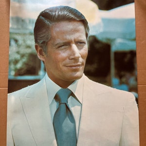 May include: A man in a white suit with a blue tie. He is smiling and looking to the right. The background is blurred and out of focus.